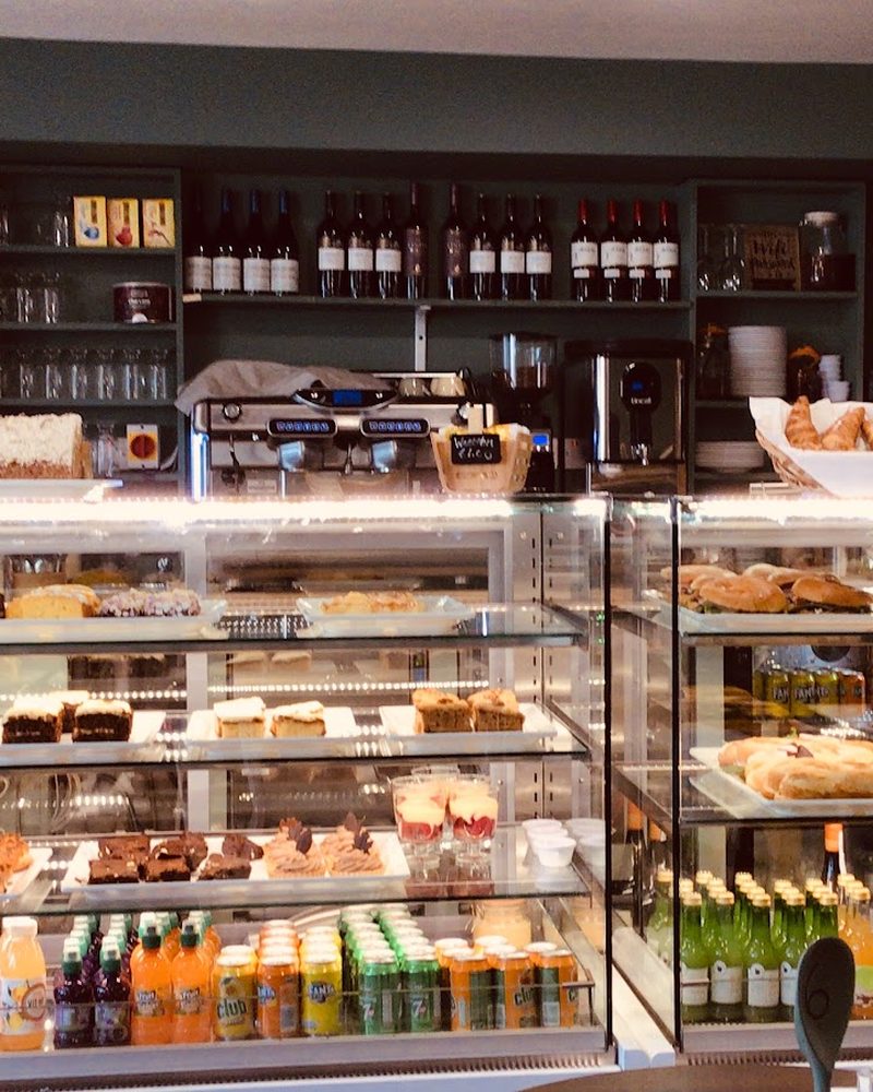 The cake and sandwich display at the Cafe Ed counter, with the coffee machine and a shelf of wine behind.